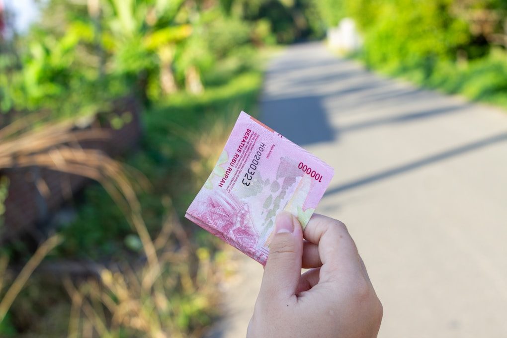 Tangan warga memegang uang seratus ribu rupiah dengan latar belakang jalanan pedesaan sebagai ilustrasi daya beli masyarakat kelas bawah saat nilai tukar rupiah ke dolar AS naik