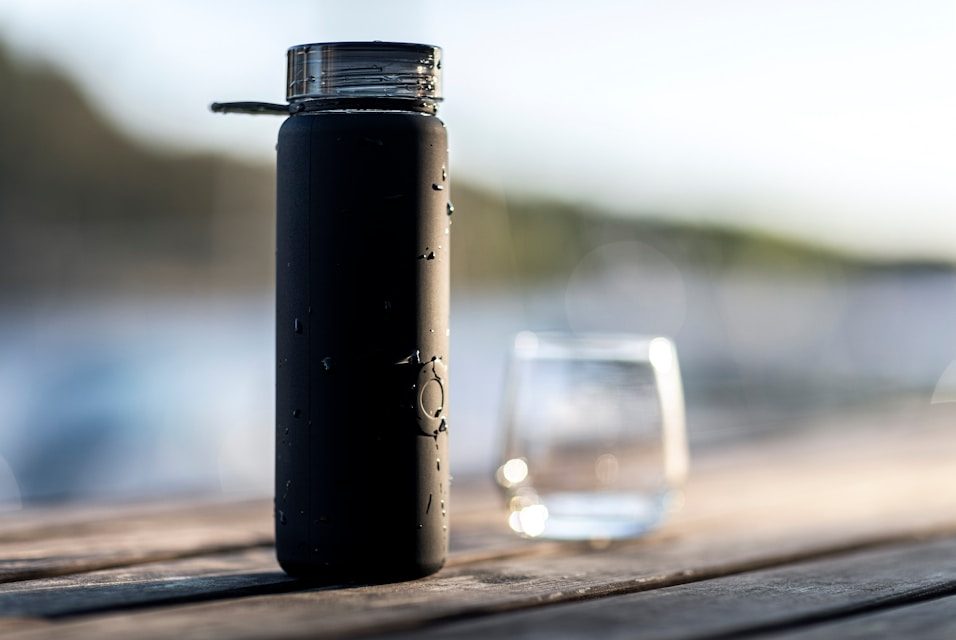 Orang yang Selalu Bawa Botol Air Punya Keunikan, Apa Saja? black bottle beside clear drinking glass on brown wooden table